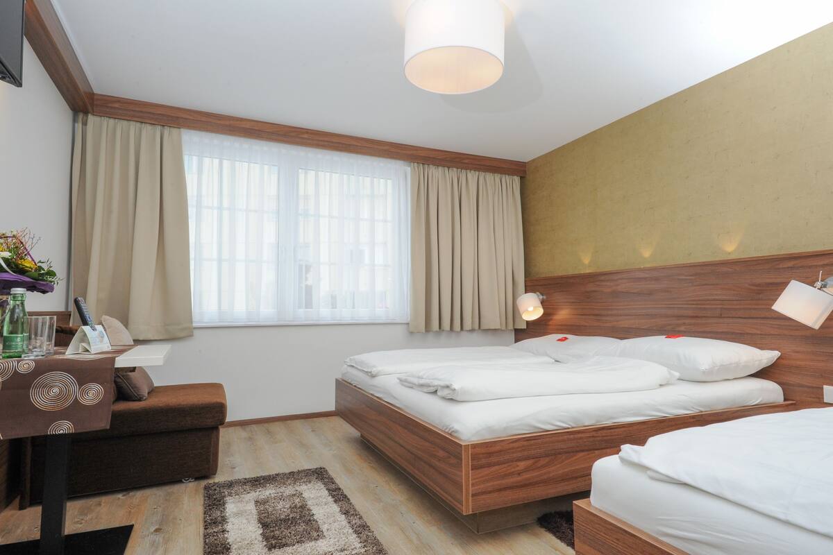A hotel room with a wooden bed, a desk with a chair, a small table with a bottle and glasses, and a window with curtains.