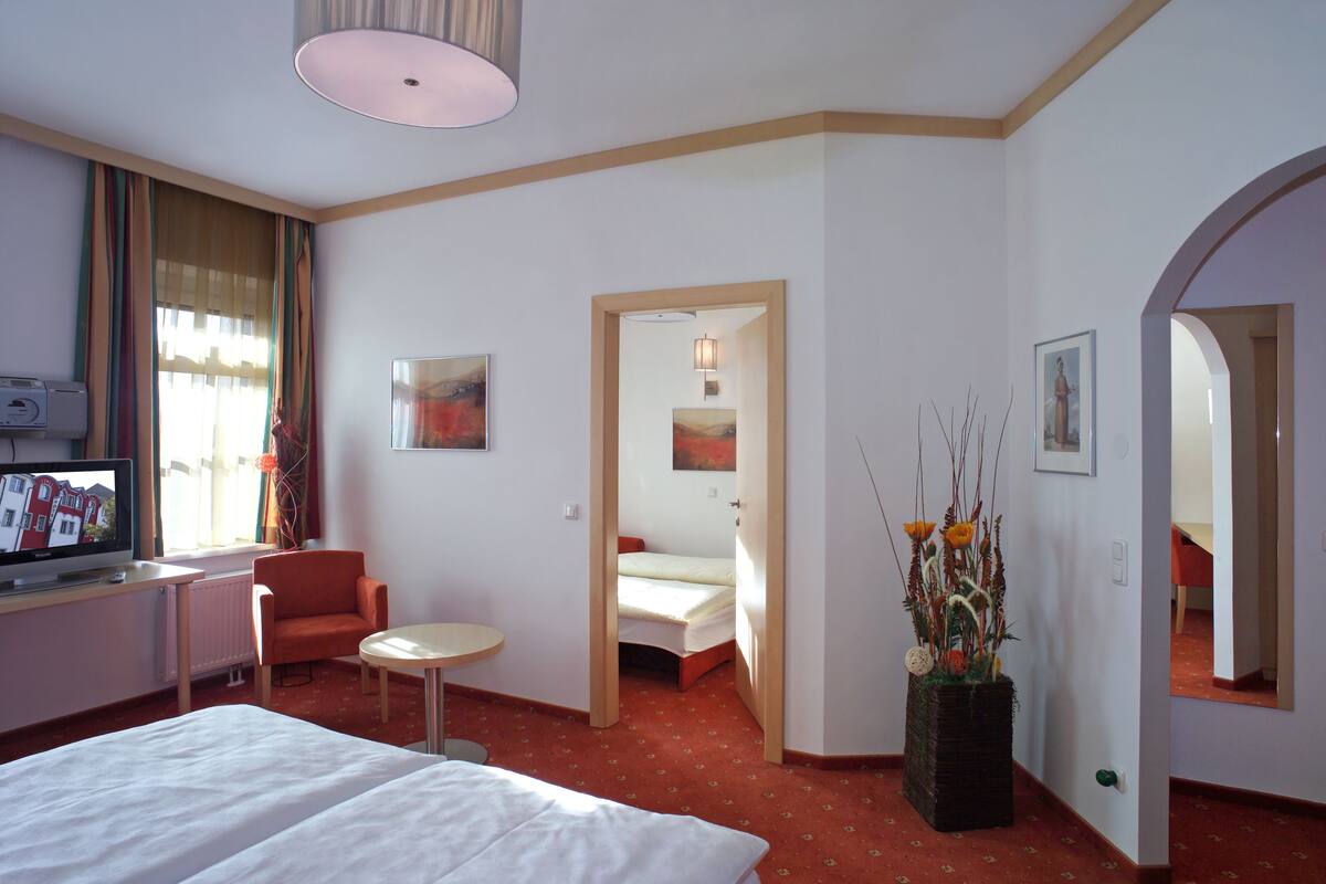 A hotel room with a bed, a desk with a computer, a chair, a small table, a potted plant, and a painting on the wall.