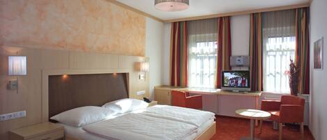 A hotel room with a bed, bedside table, lamp, TV, desk, chair, and a small table.