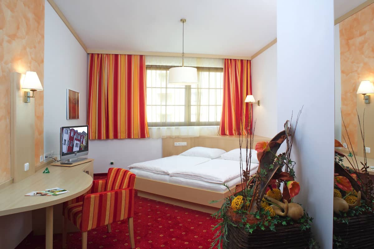 A hotel room with a bed, a desk with a computer, a chair, a window with curtains, and a decorative plant.