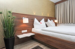 A modern hotel room with a wooden headboard, a bedside table, a potted plant, and a bed with white bedding.