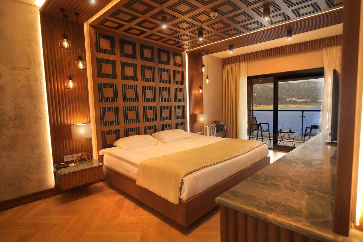 Quarto de hotel com uma cama grande, painel de parede de madeira e vista para um lago.