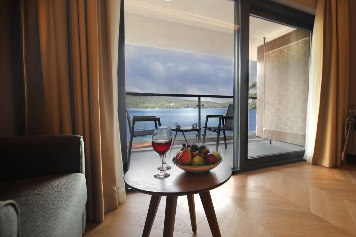 Quarto com vista para um lago, uma mesa com uma cesta de frutas e uma taça de vinho tinto, e uma cadeira.
