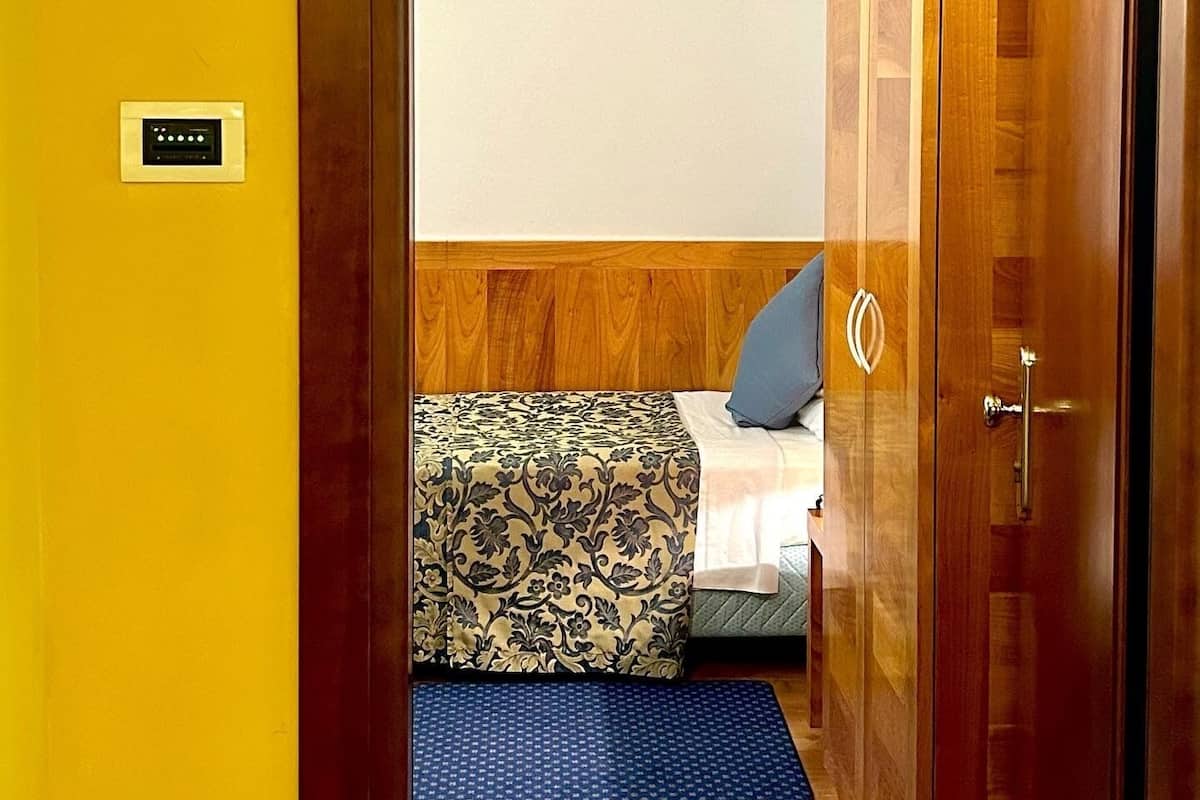 A hotel room with a bed, a bedside table, a nightstand, a wall-mounted control panel, and a door with a wooden finish.