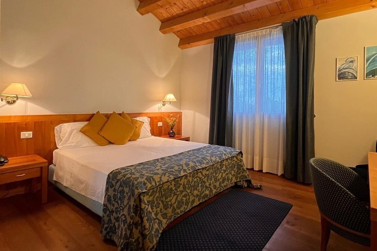 A hotel room with a wooden ceiling, a bed with a floral bedspread, a nightstand with a lamp, a chair, and a window with curtains.