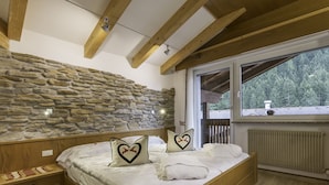A bedroom with a stone wall, wooden beams, a large window, and a bed with heart-shaped pillows. - Hotel Ramon (Campitello di Fassa)
