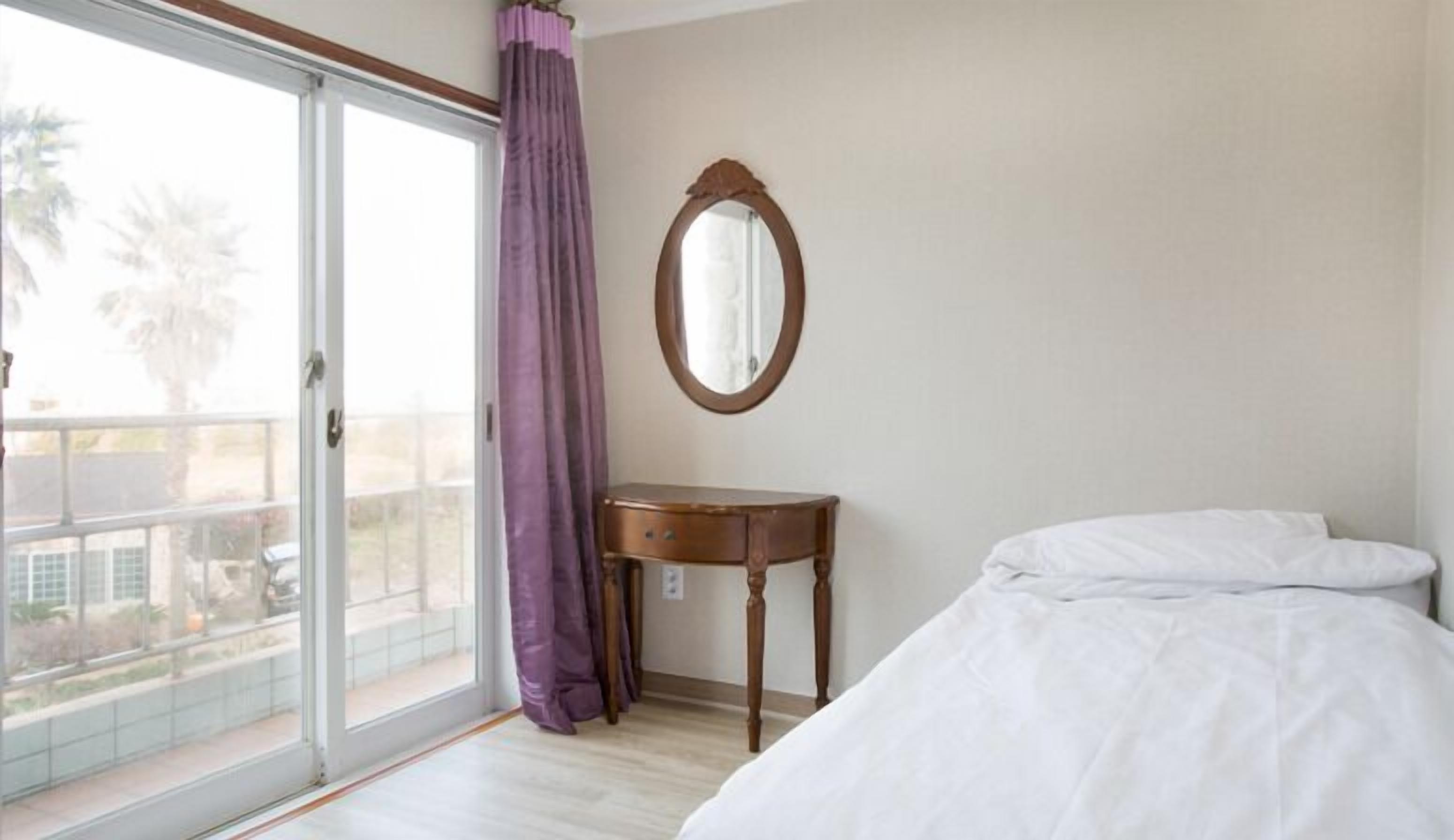 A bedroom with a bed, a small wooden nightstand, a mirror, and a balcony with purple curtains.