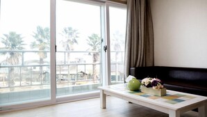 Una habitación con puerta corrediza de vidrio que da a un balcón con palmeras, un sofá negro y una mesita con un florero y un libro.
