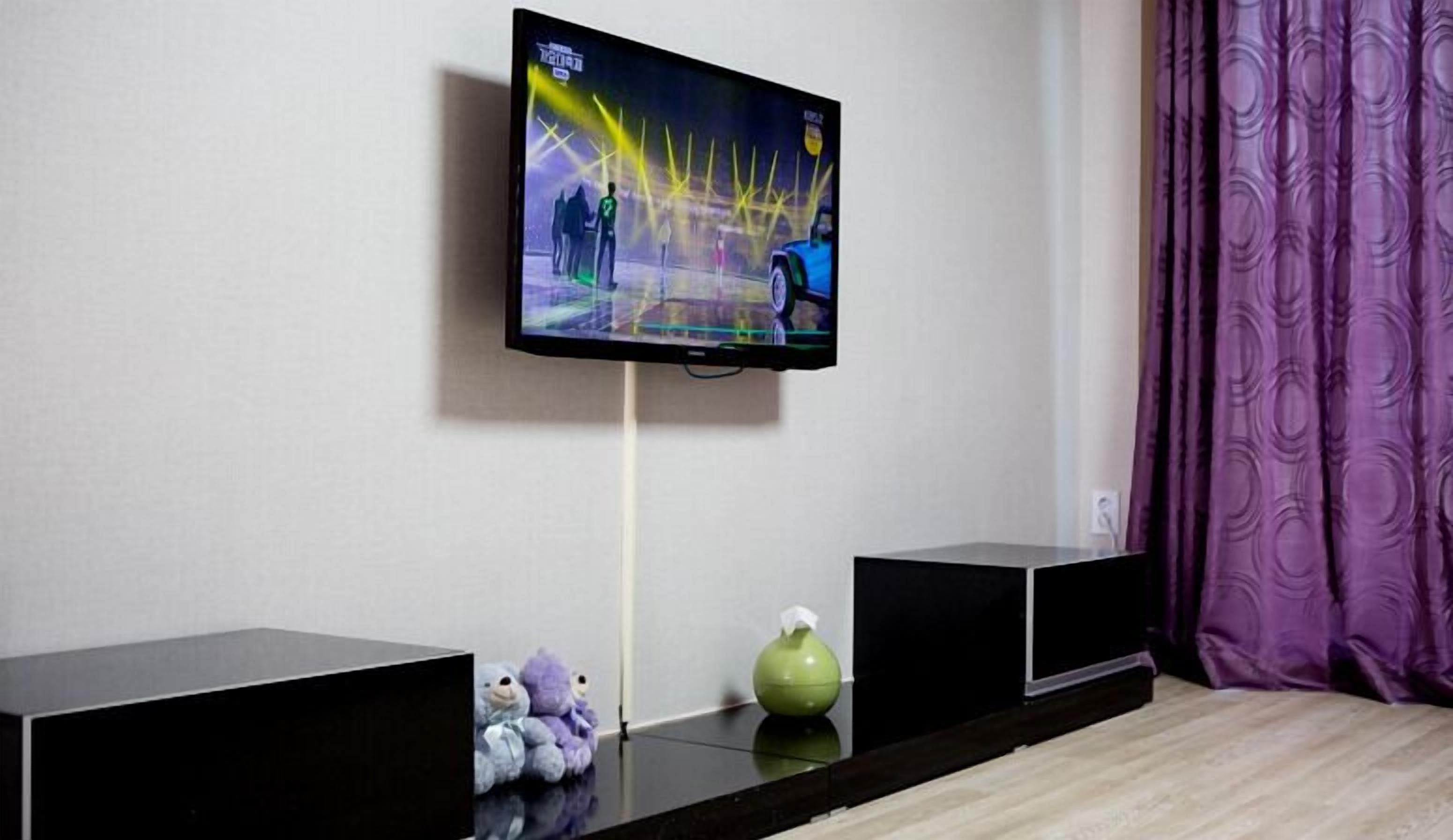 A flat-screen TV mounted on a wall, two black speakers, a green vase, and a purple curtain.
