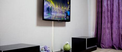 A flat-screen TV mounted on a wall, two black speakers, a green vase, and a purple curtain.