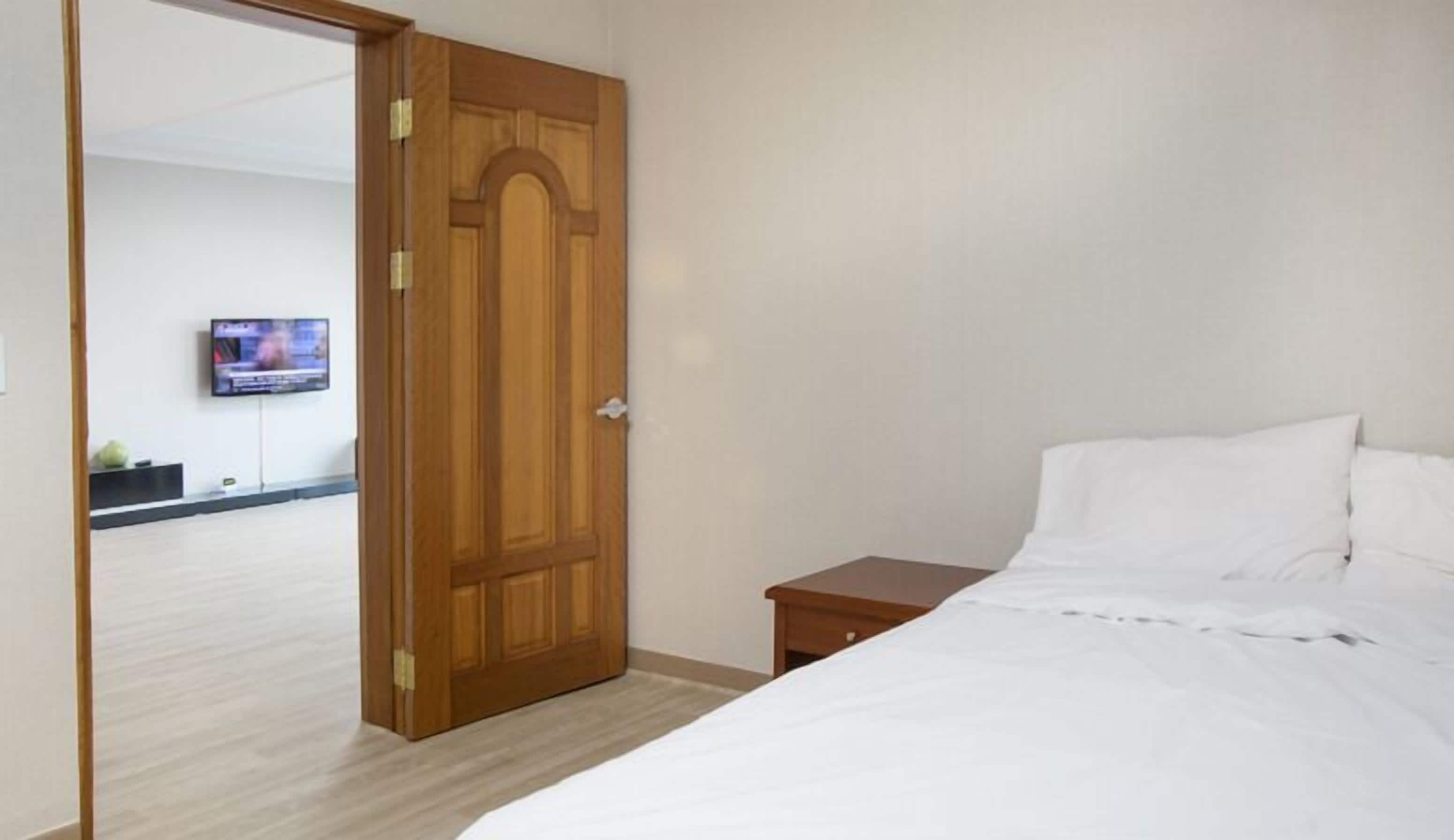 A hotel room with a wooden door, a bed with white linens, a small wooden nightstand, and a television mounted on the wall.