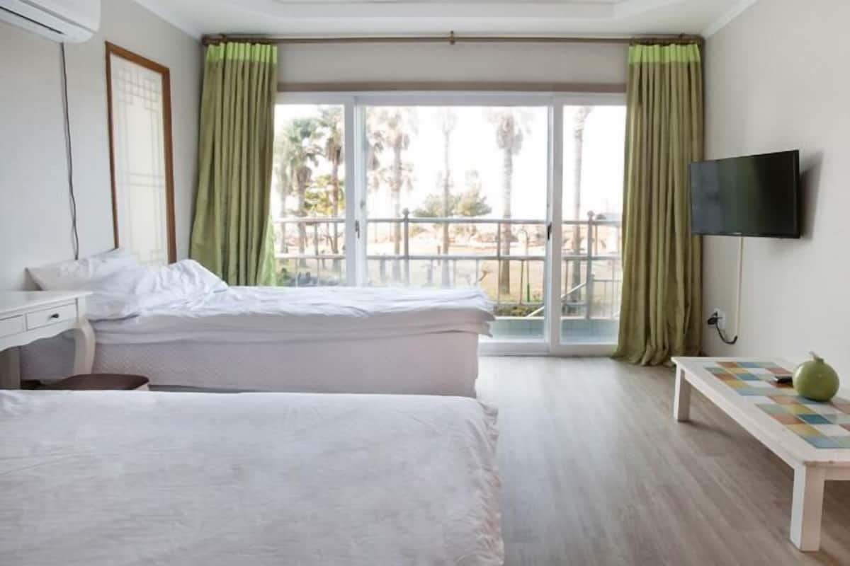 A hotel room with two beds, a desk, a television, and a balcony with a view of palm trees.