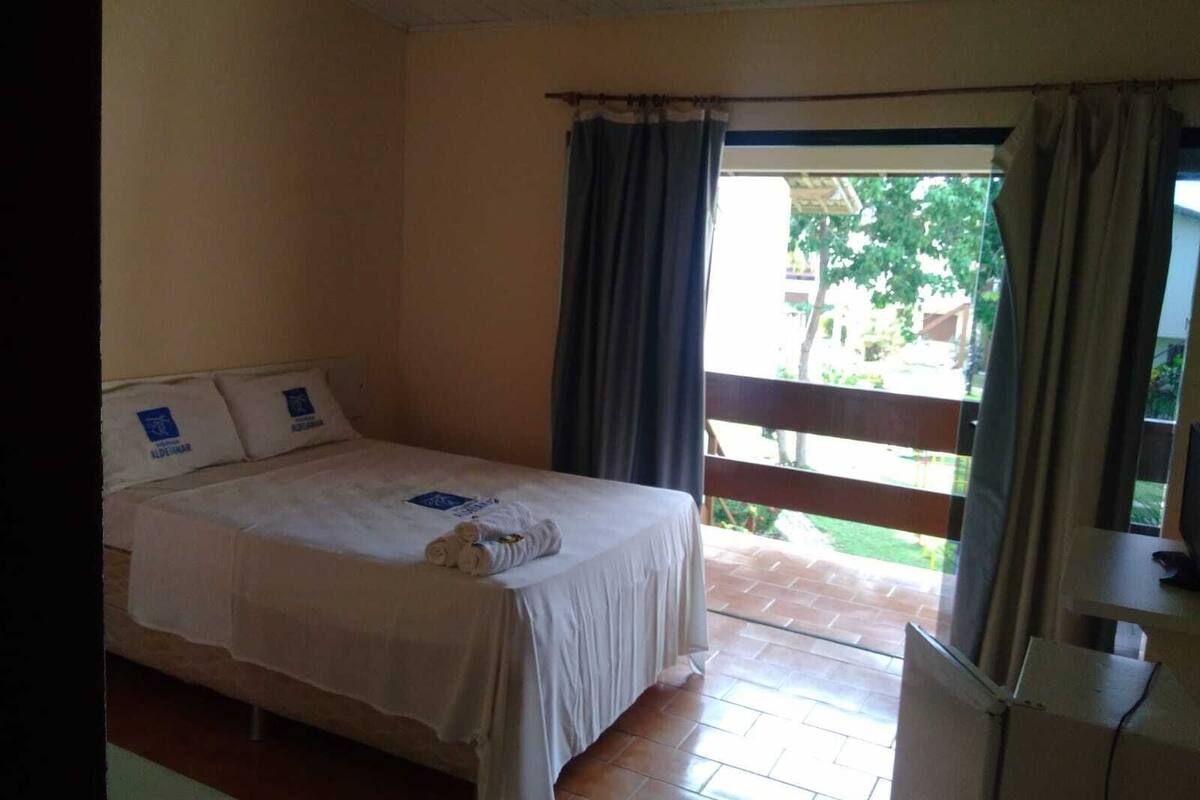 A single bed with a white bedspread and pillows, a small table with a chair, and a window with curtains.