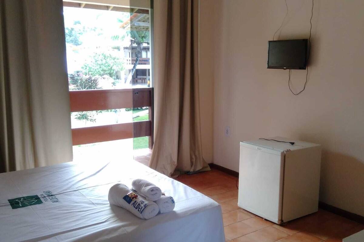 A hotel room with a bed, a small white refrigerator, a TV mounted on the wall, and a view of the outside through a window.