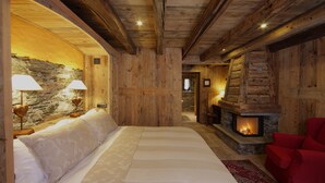 Ein Interieur einer Blockhütte mit einer Steinwand, Holzbalken, einem Kamin, einem Bett mit weißer Bettwäsche und einem roten Sessel.