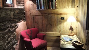 Ein gemütliches Zimmer mit einem roten Sessel, Holzregalen mit Büchern, einer Lampe und einer Steinsäule.