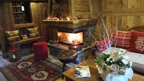 Ein gemütlicher Raum mit Kamin aus Stein, Holzwänden, einer roten Bank, einem gemusterten Teppich und einem Tisch mit Buch und Blumen.