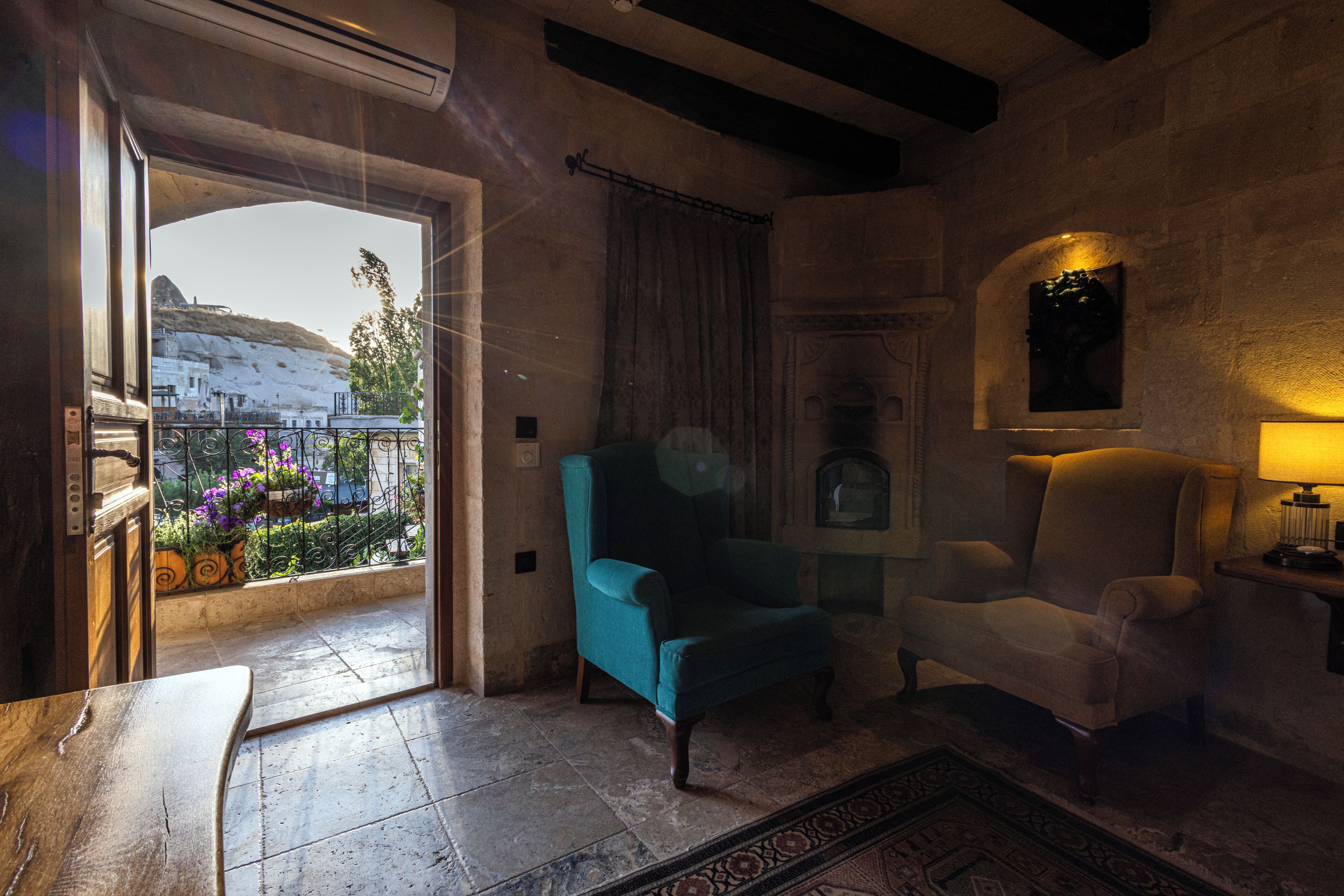 A room with a stone wall, a blue armchair, a white armchair, and a view of a balcony with flowers.
