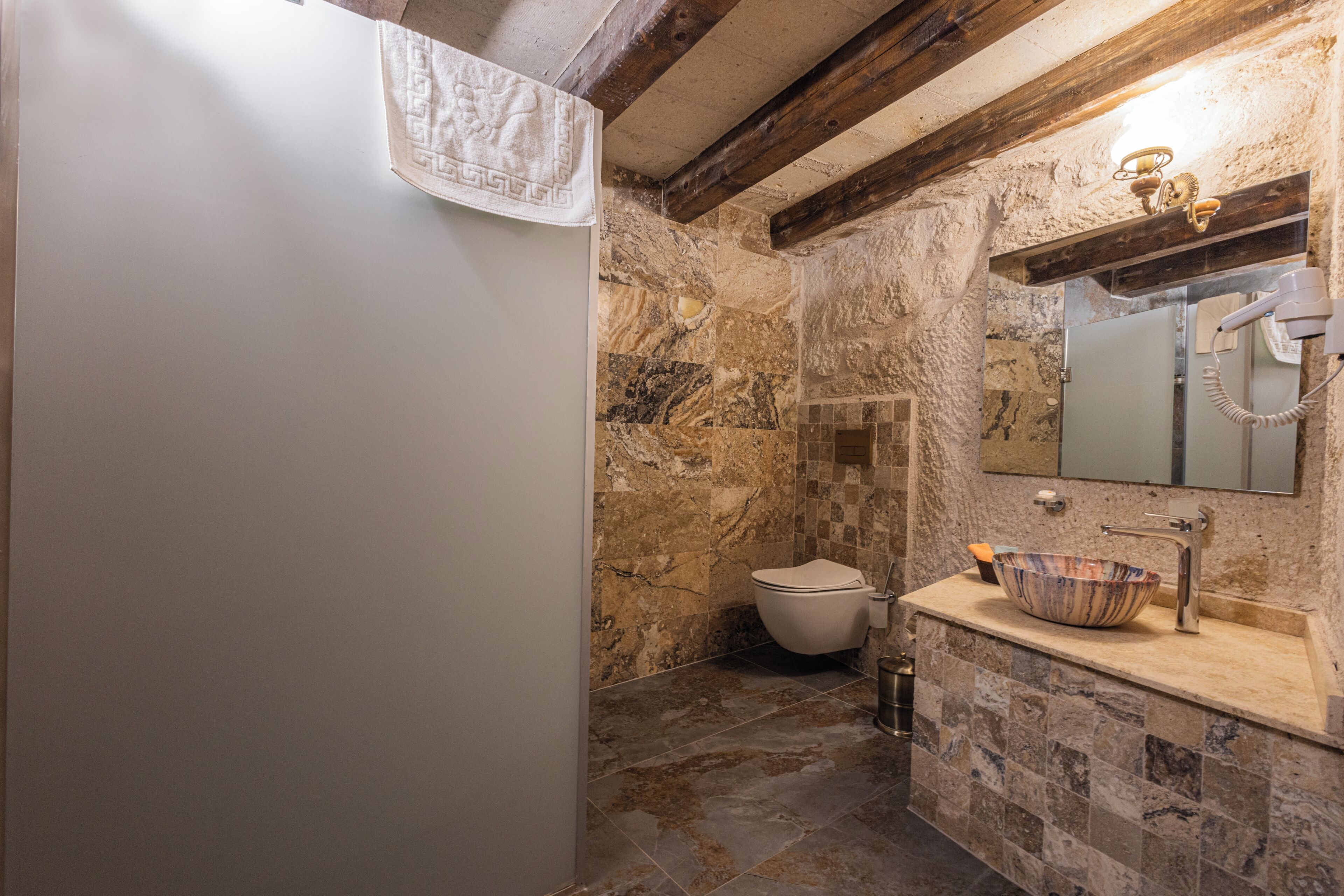 Une salle de bain avec un mur en pierre, des toilettes blanches, un lavabo avec un receveur en marbre et une serviette accrochée au mur.