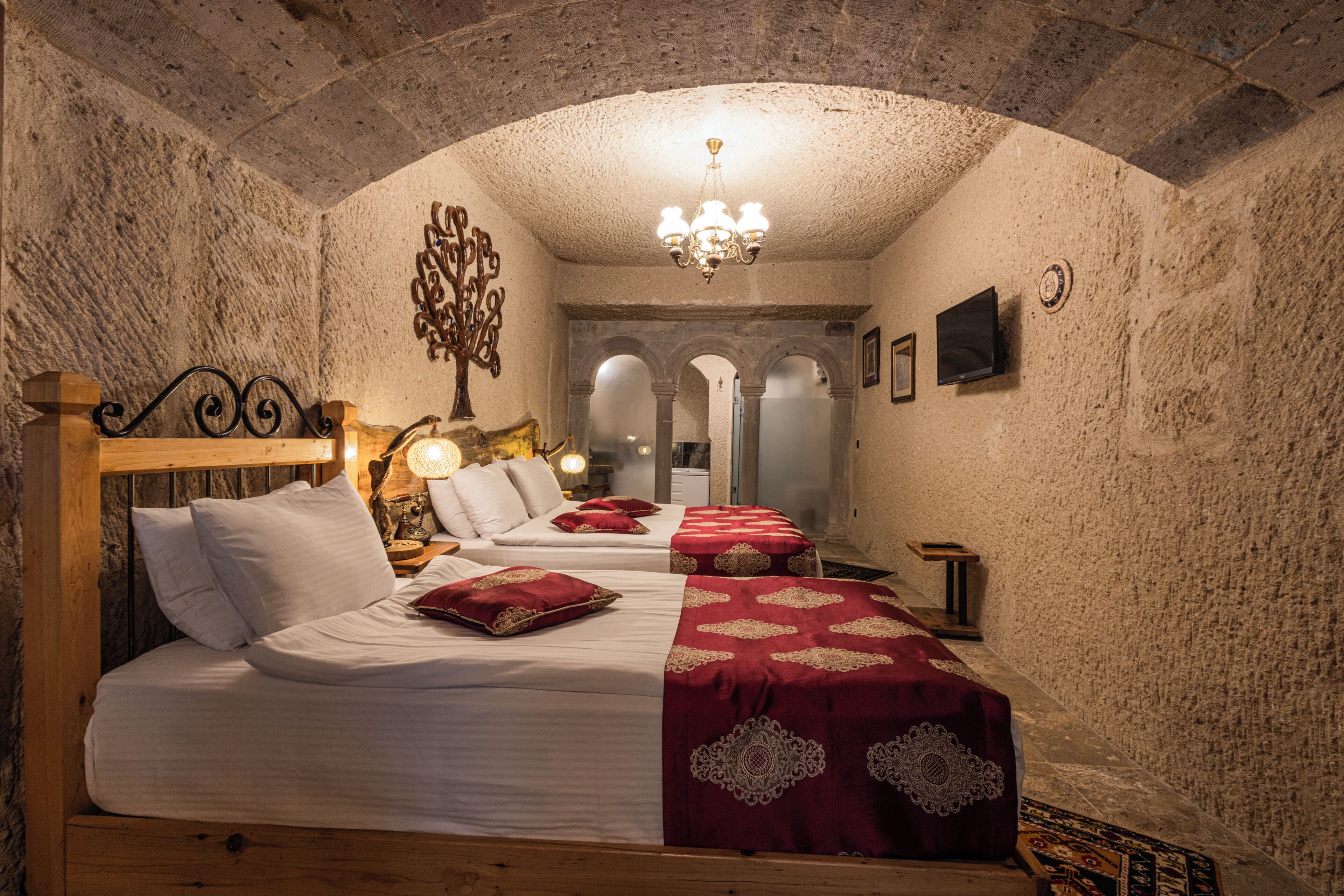 A room with a vaulted ceiling, a bed with white linens and red pillows, a chandelier, and a television on the wall.