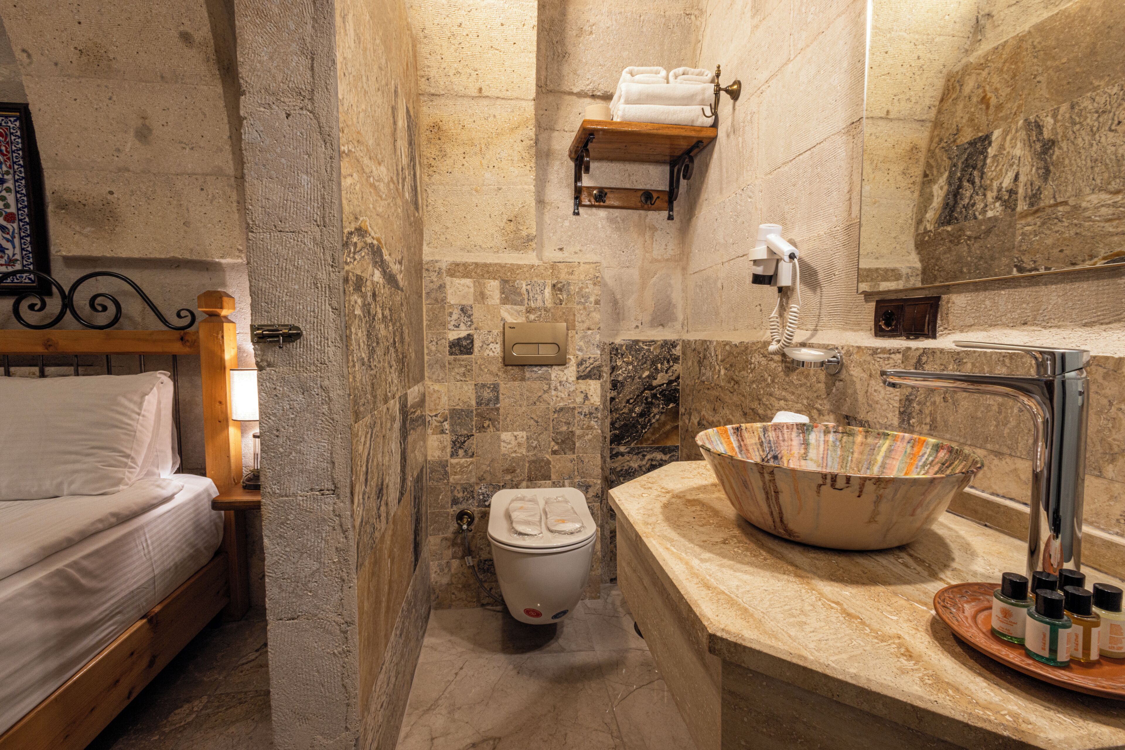 A bathroom with a stone wall, a wooden bed, a white toilet, a marble sink, and a mirror.