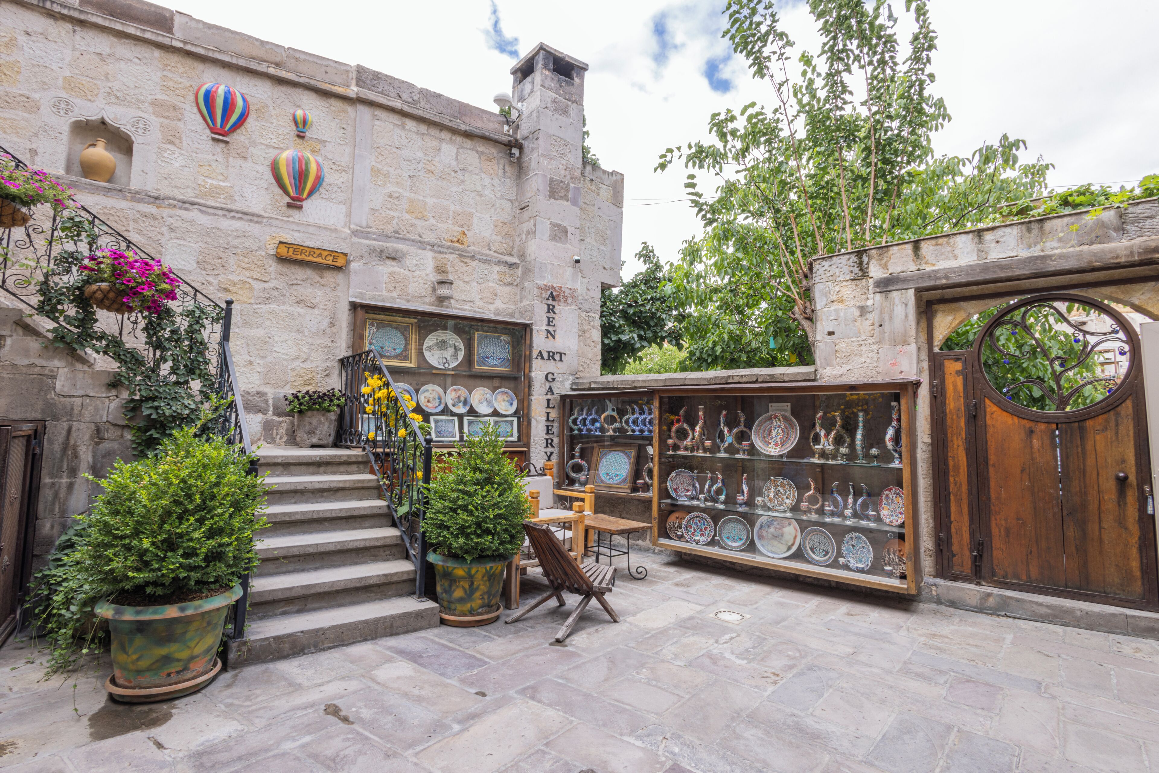 A courtyard with stone steps leading to a terrace, surrounded by potted plants and a shop displaying ceramic plates.