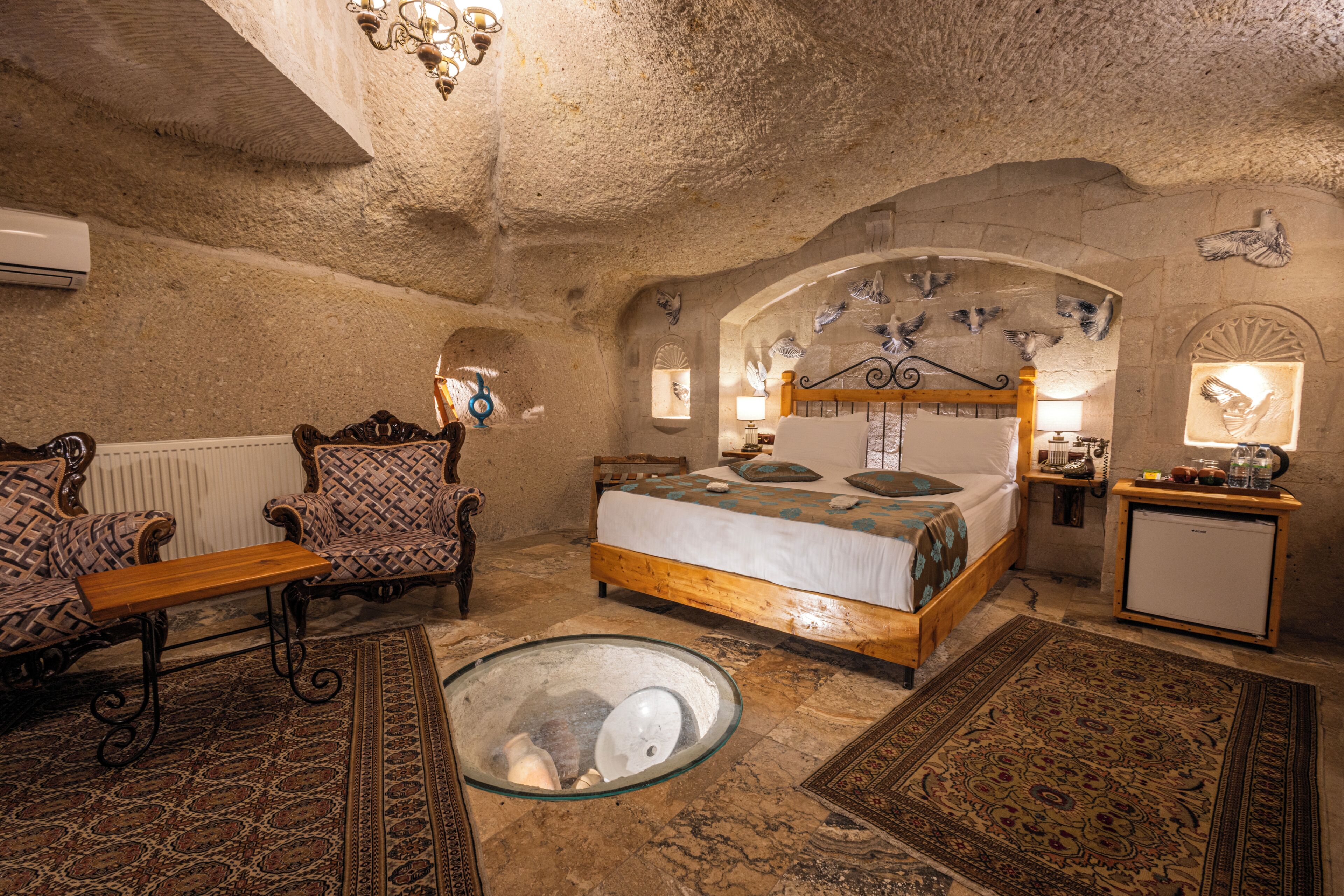 A room with a stone wall, a bed with a wooden headboard, a small table, a chair, and a glass table with a white object.