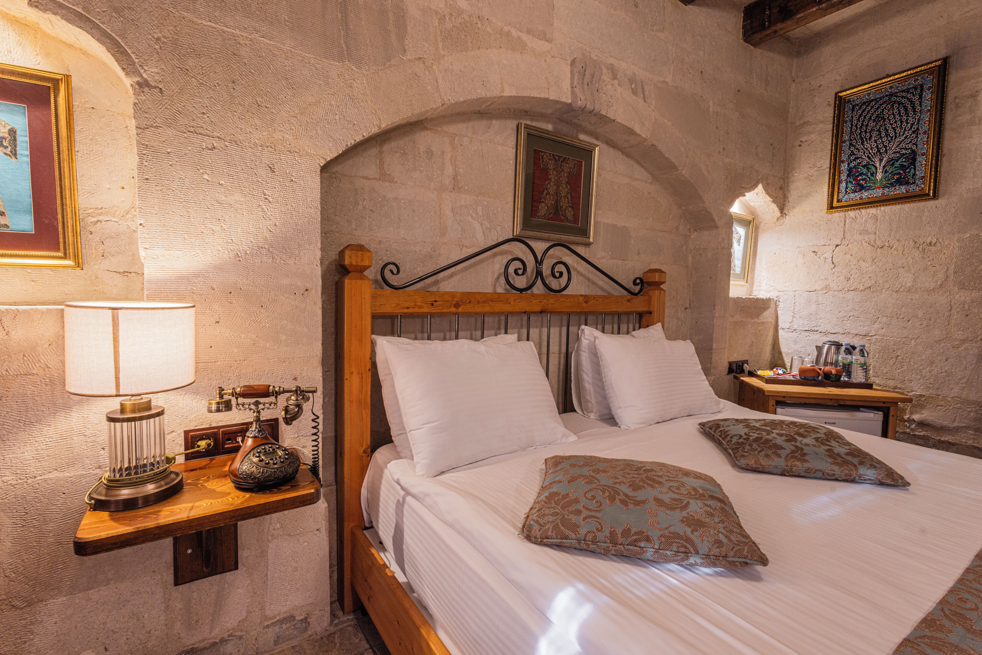 Una habitación con pared de piedra, cama de madera con ropa de cama blanca, teléfono antiguo y una mesita con una lámpara.