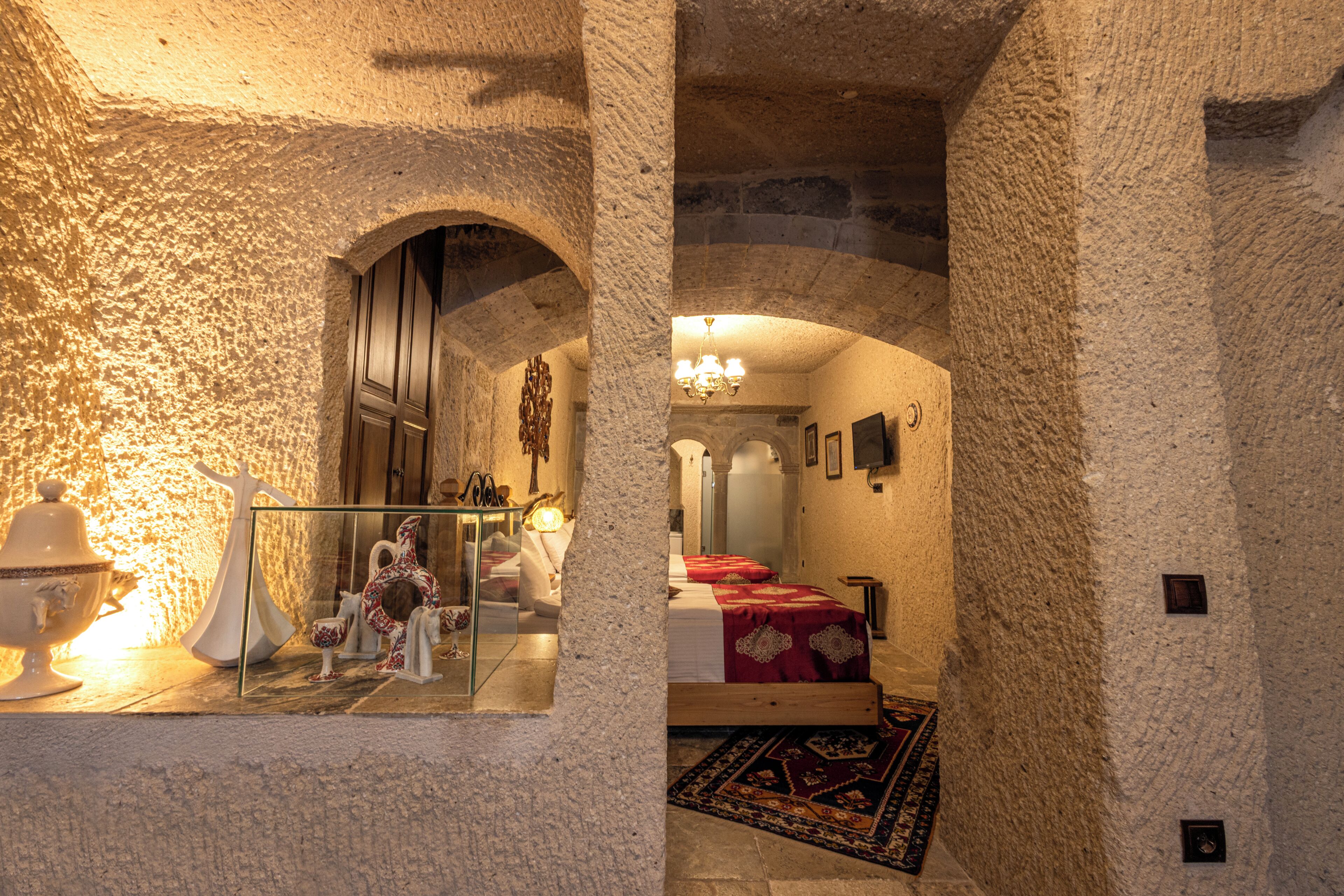 A room with stone walls, a bed with red bedding, a chandelier, and a mirror reflecting the room.
