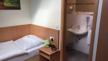Une petite chambre à coucher, bien rangée, avec un lit simple, une table de chevet avec une lampe et un miroir fixé au mur dans la salle de bain attenante.