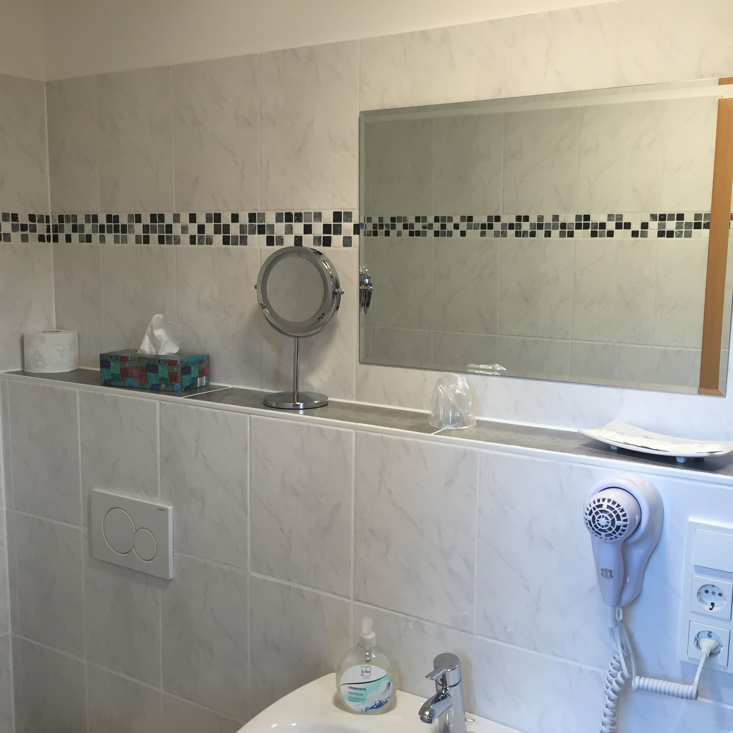A bathroom with a toilet, a sink, a mirror, a hairdryer, and a soap dispenser.