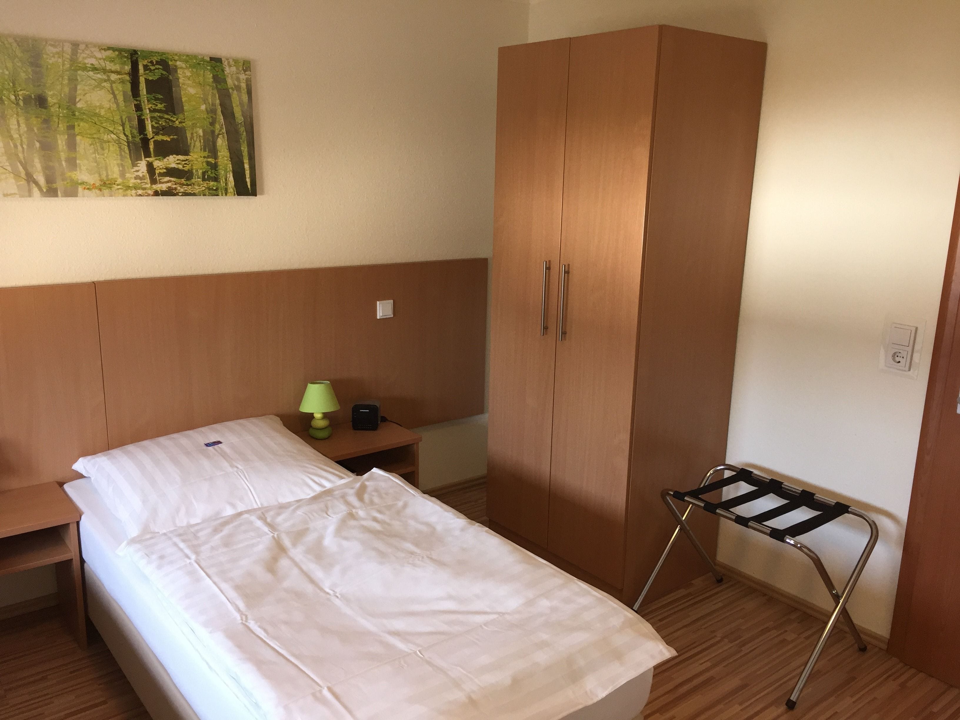 A small, neatly arranged bedroom with a bed, a nightstand, a lamp, a wardrobe, and a drying rack.