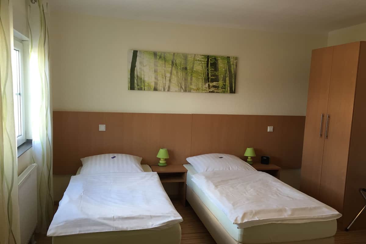 A room with two single beds, a nightstand with a lamp, a wardrobe, and a painting on the wall.