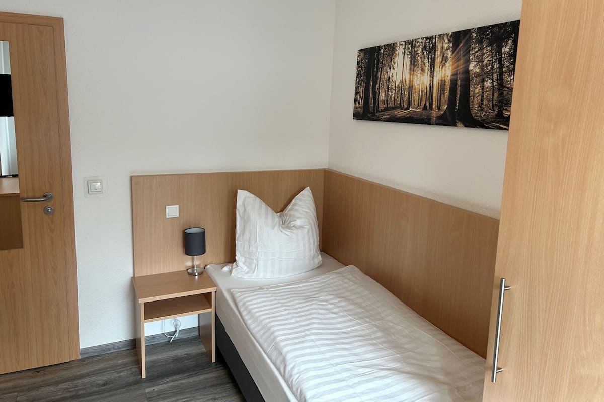 A small, neatly arranged bedroom with a single bed, a nightstand, a lamp, and a painting of a forest on the wall.