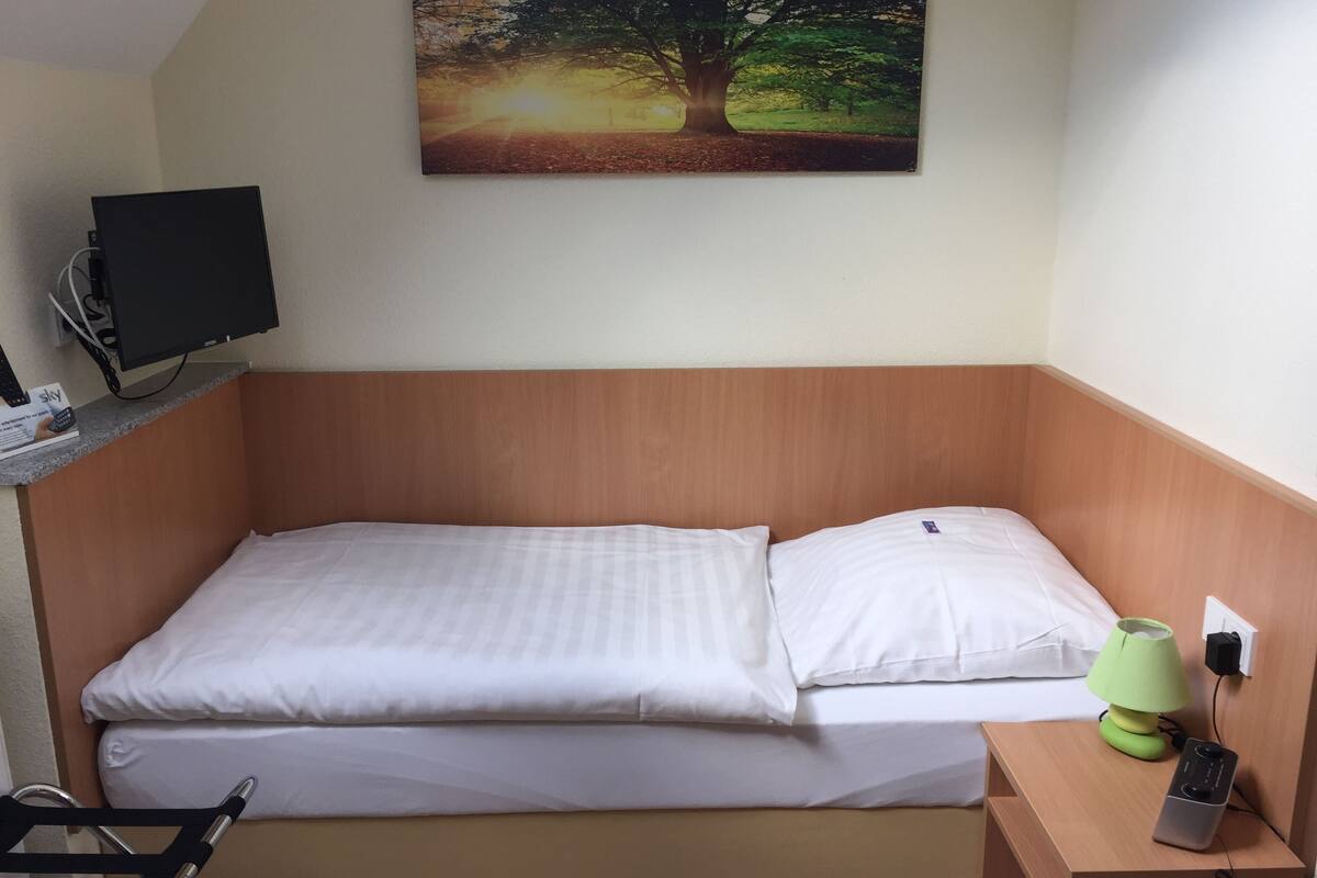 A single bed with white linens, a wooden headboard, a desk with a computer monitor, a green desk lamp, and a power strip.