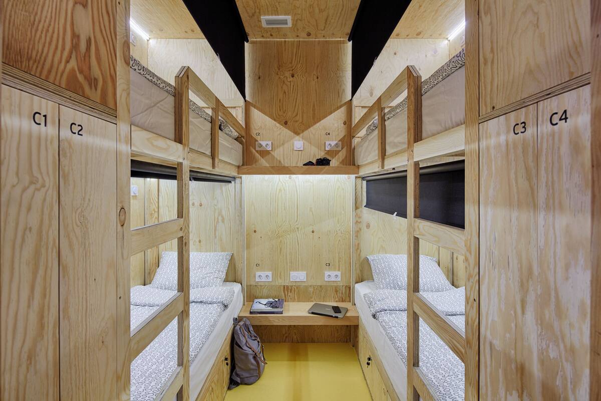 Une petite pièce de style cabane en bois, avec des lits superposés numérotés de C1 à C4, une petite table avec un ordinateur portable et un sac à dos, et un sol jaune.