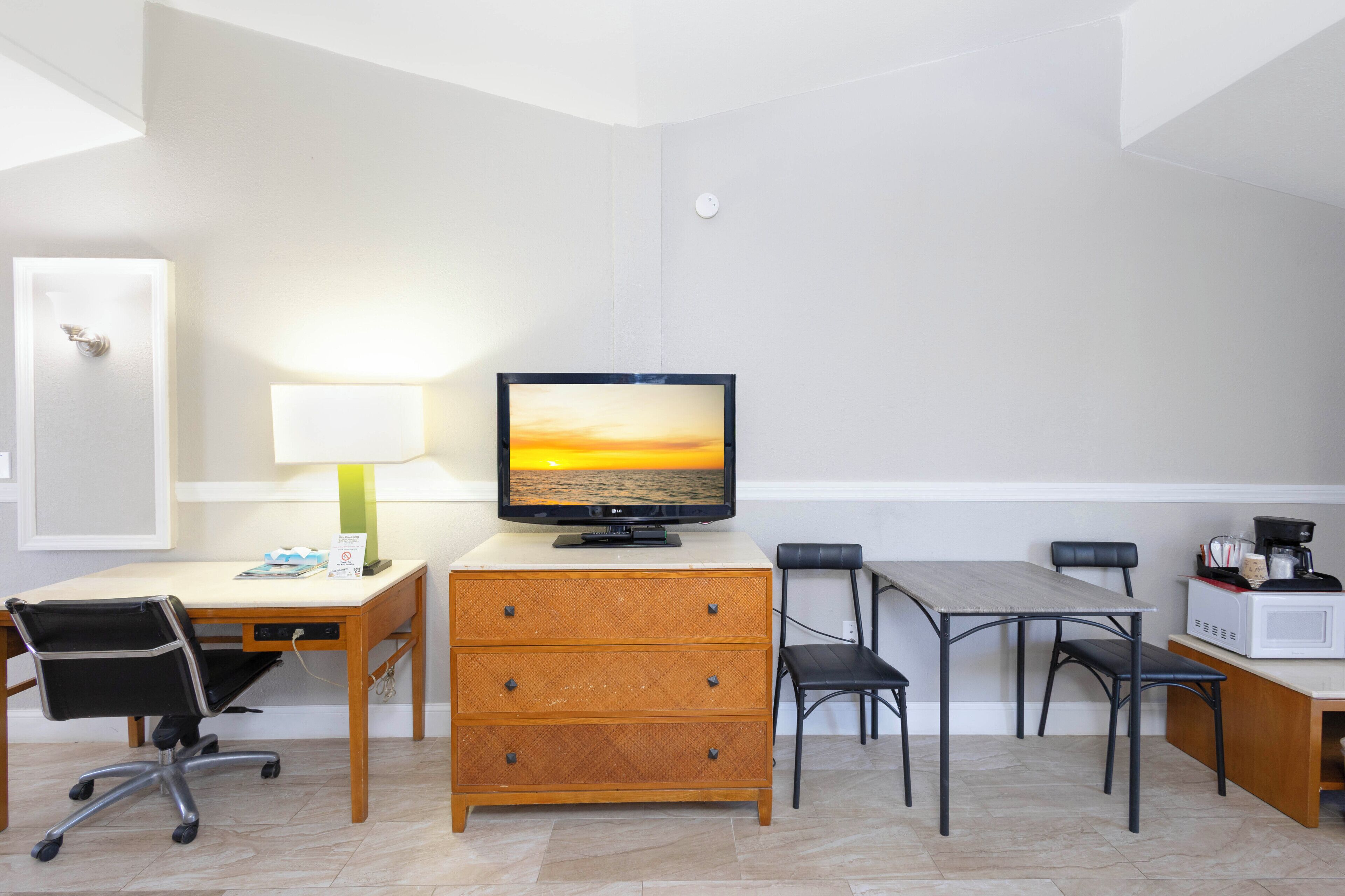 Habitación de hotel con un escritorio de madera, un televisor mostrando un atardecer, una mesa de comedor con sillas, un microondas y una cafetera.