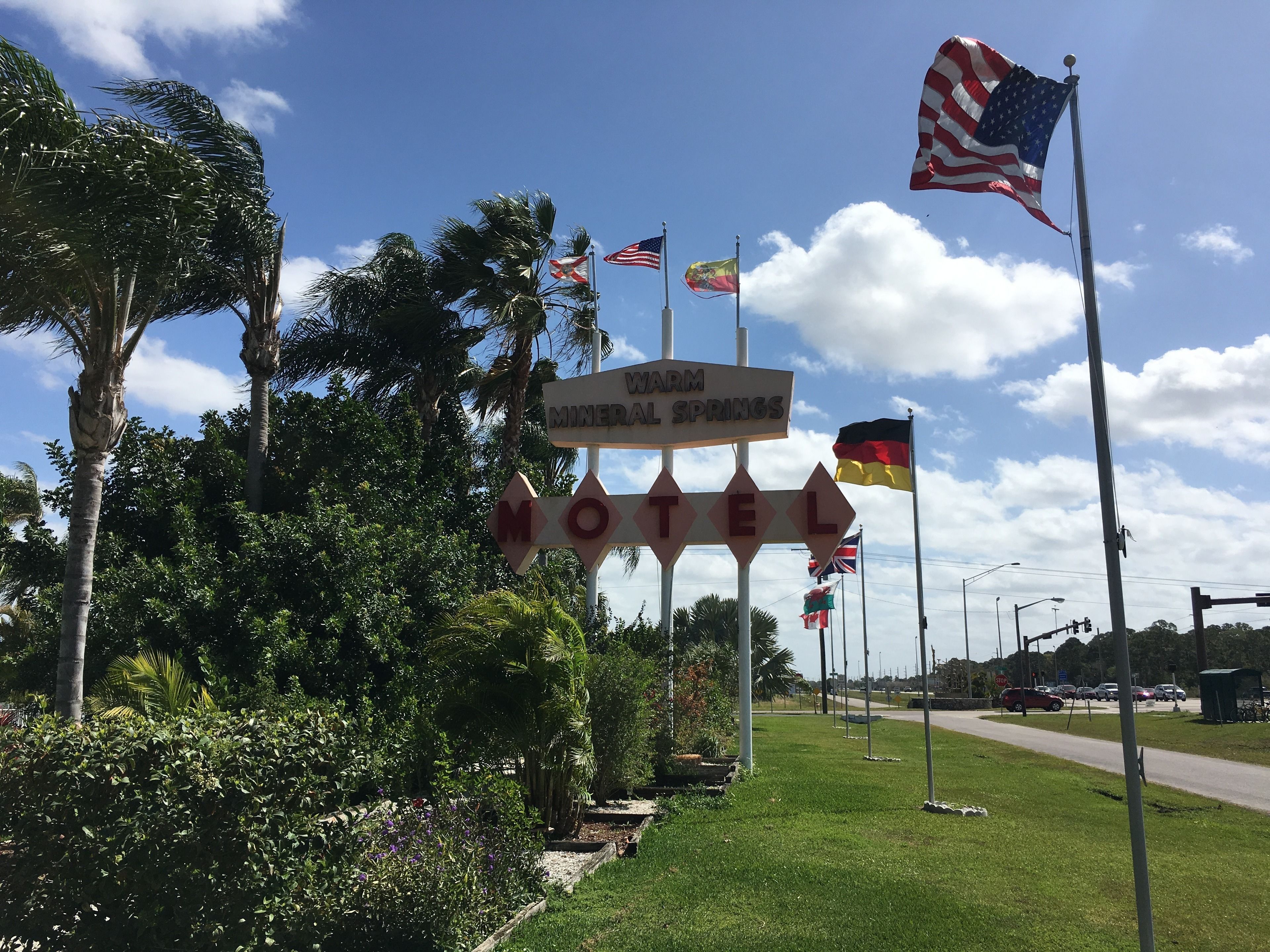 A sign for the Warm Mineral Springs Hotel with multiple flags, including the American flag, and surrounded by lush greenery.