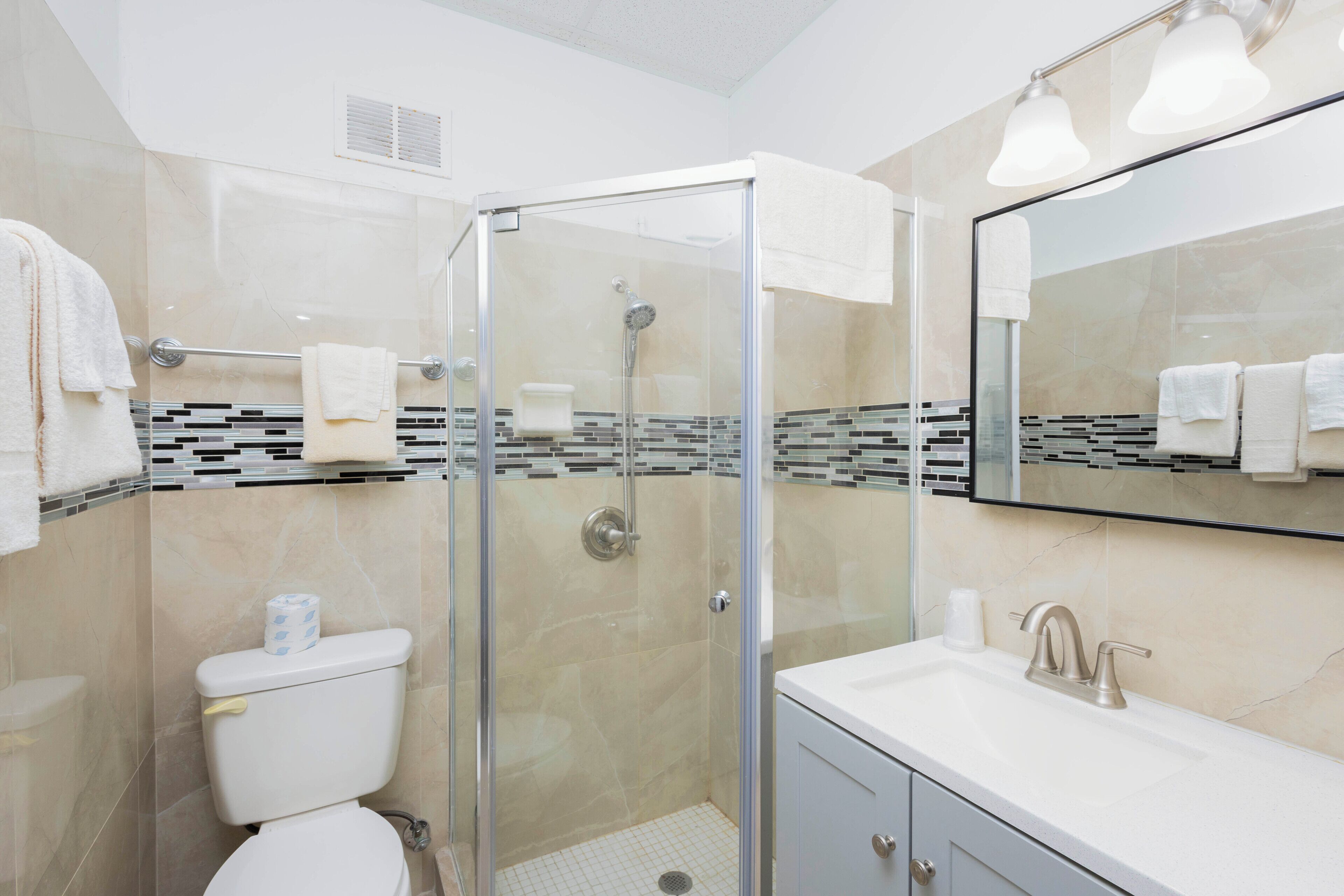 Une salle de bain moderne dotée d’une douche à parois de verre, de toilettes, d’un lavabo avec un miroir et d’un mur recouvert de carreaux.