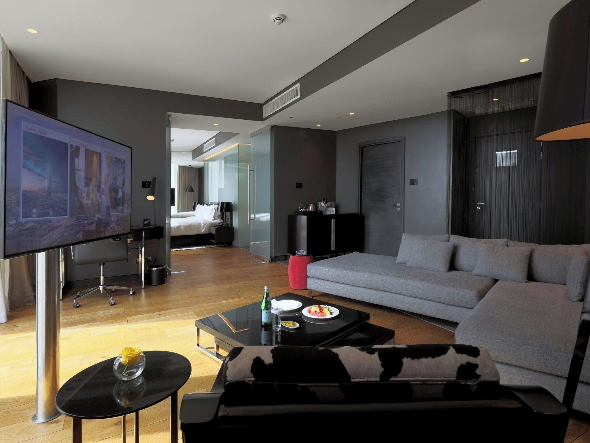 A modern living room with a large flat-screen TV, a sofa, a coffee table with food, and a bed in the background.