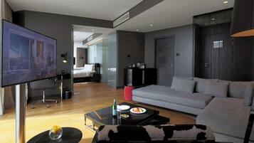 A modern living room with a large flat-screen TV, a sofa, a coffee table with food, and a bed in the background.