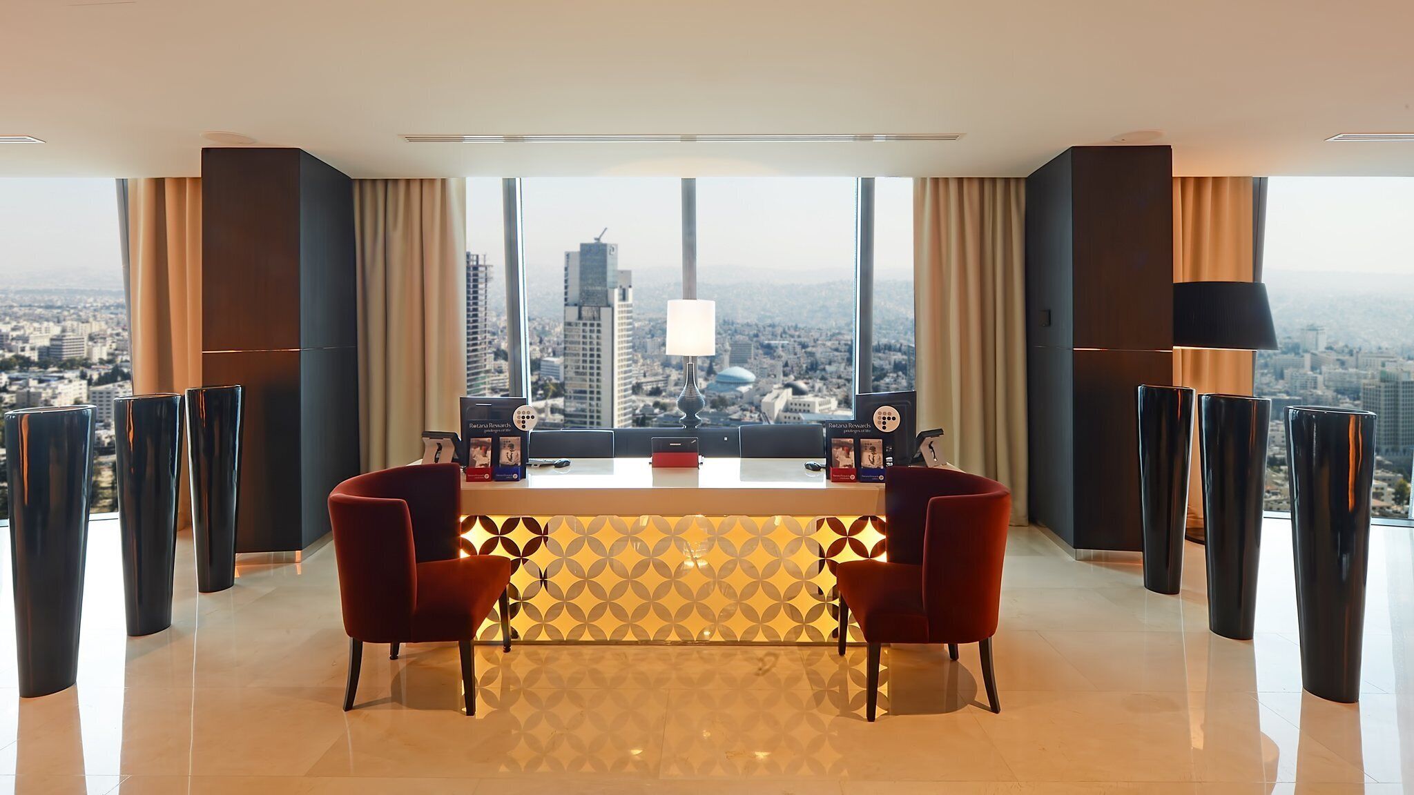 A modern hotel room with a city view, a dining area with two red chairs, a patterned rug, and a large window with curtains.