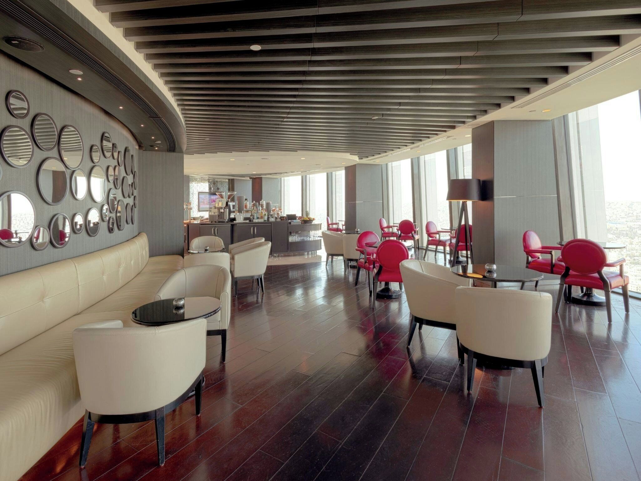 A modern hotel lobby with a curved ceiling, a seating area with white and red chairs, a bar, and a wall with circular mirrors.