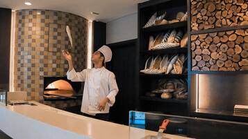 Un chef con uniforme y gorro blanco de pie en una cocina moderna con horno de pizza, estantes con pan y un salpicadero de azulejos.