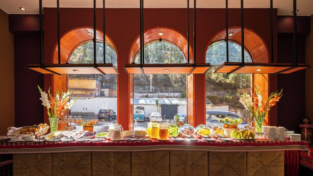 A buffet table with various food items and drinks, set against a backdrop of large arched windows overlooking a street scene. - Hotel Mamasara (Cusco)