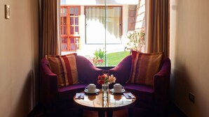 A room with a sofa facing a window, a small table with two cups and a vase of flowers, and a view of a building with a balcony. - Hotel Mamasara (Cusco)