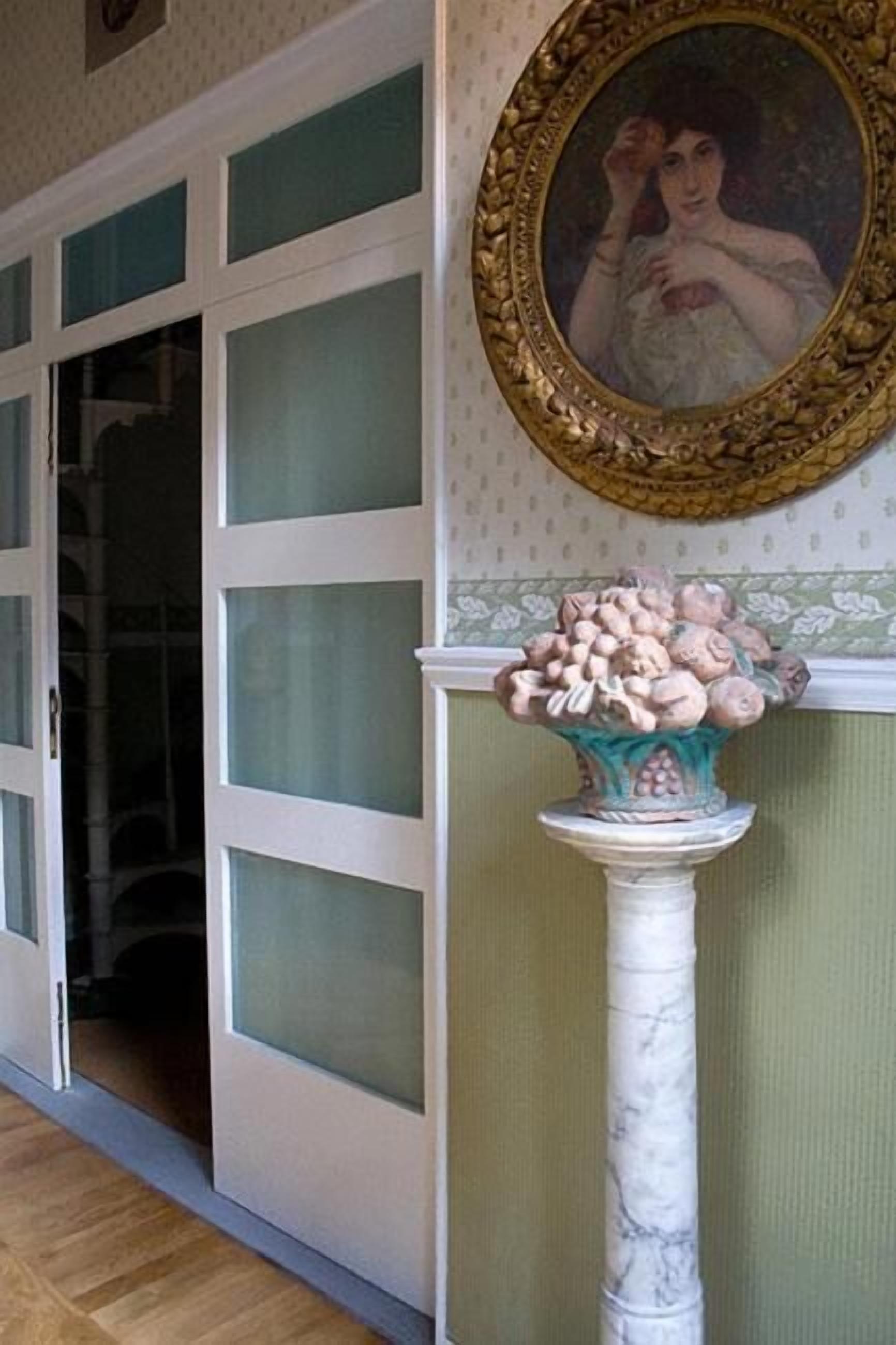 A room with a large glass door, a marble column with a decorative bowl, and a framed portrait on the wall.