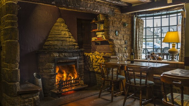 Una chimenea de piedra con fuego, sillas de madera, una mesa con copas de vino y una ventana con vista a la calle.