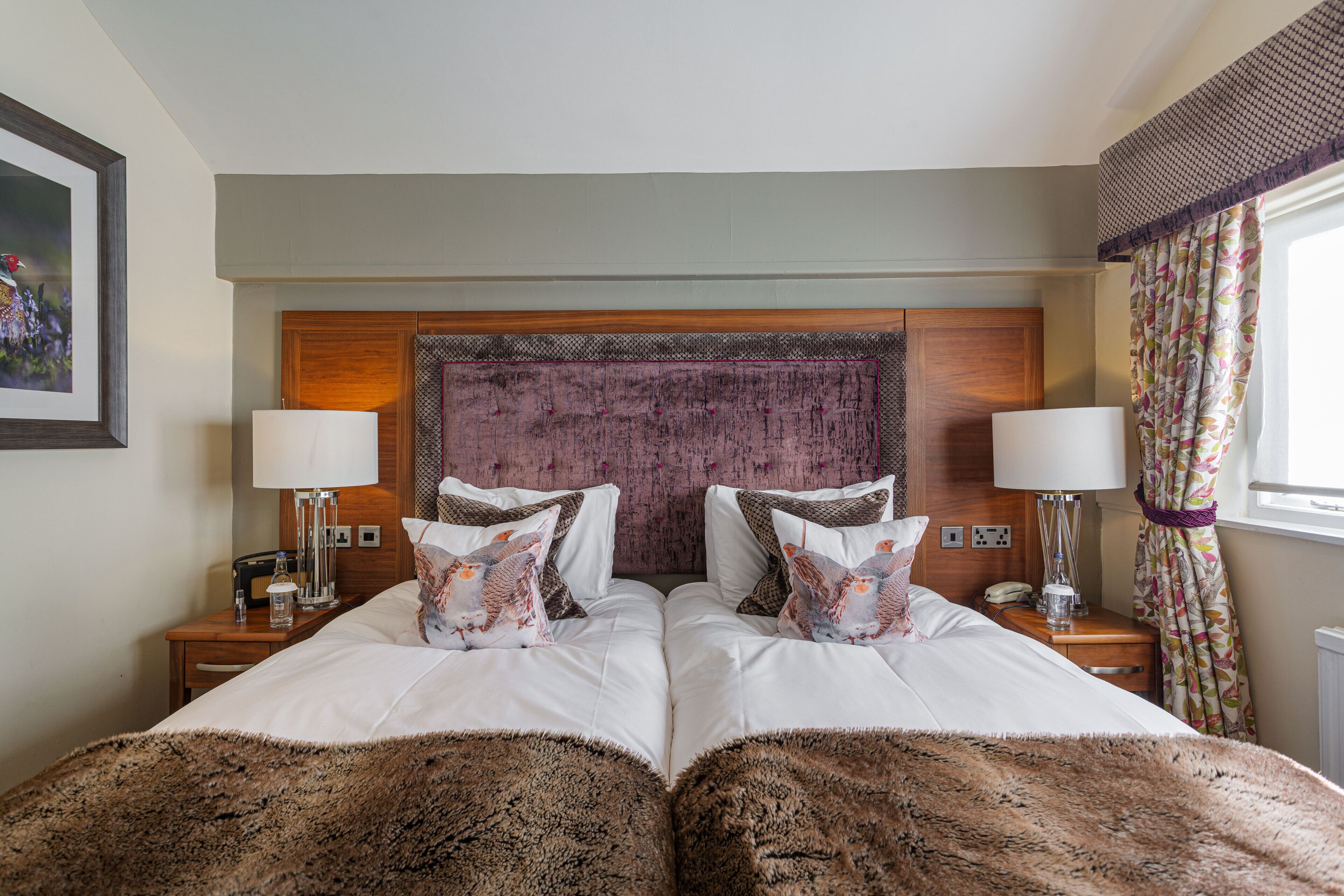 A double bed with a headboard, two bedside tables with lamps, a wooden nightstand, and a window with curtains.