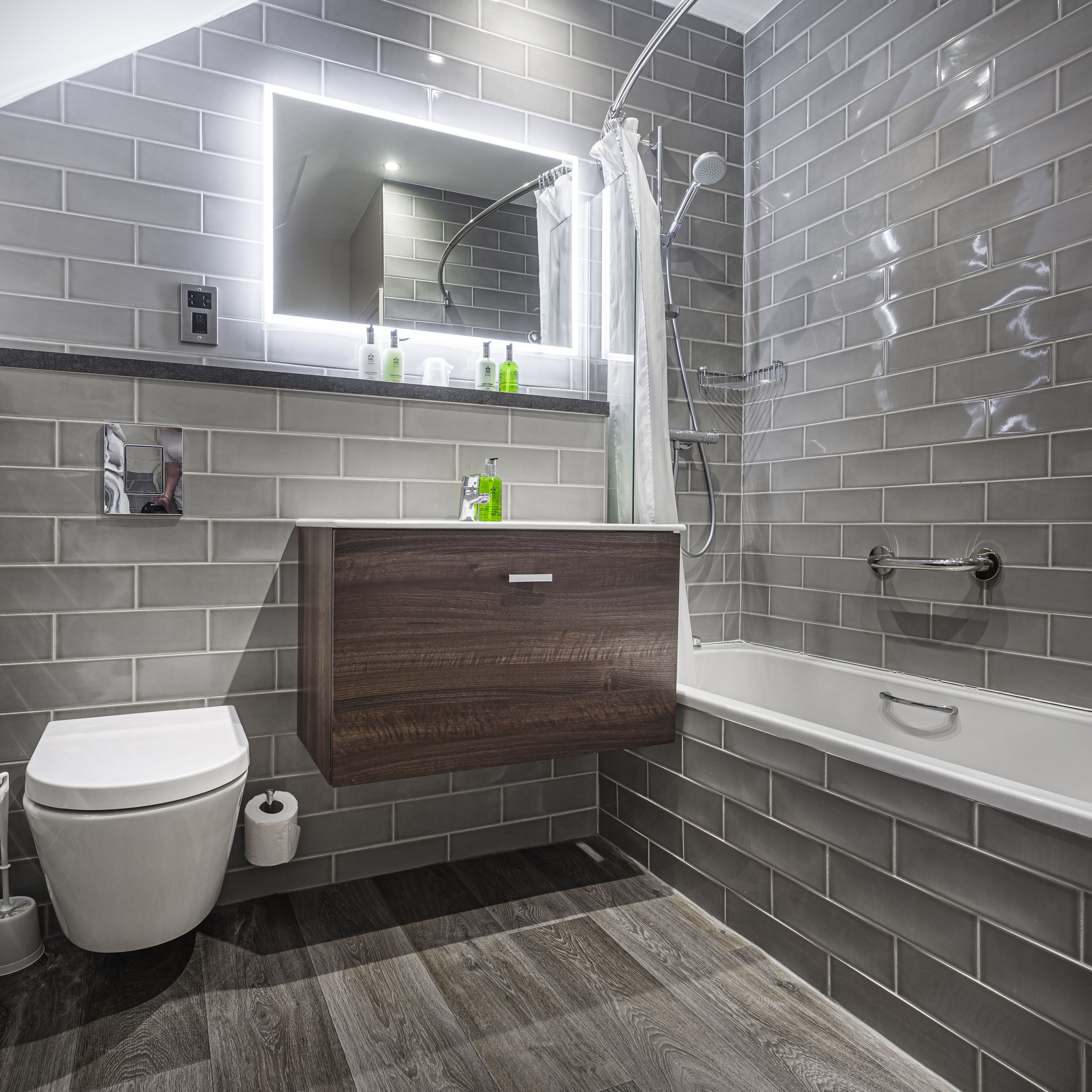 Une salle de bain moderne avec des murs recouverts de carreaux gris, un meuble-lavabo en bois, des toilettes blanches et une baignoire.